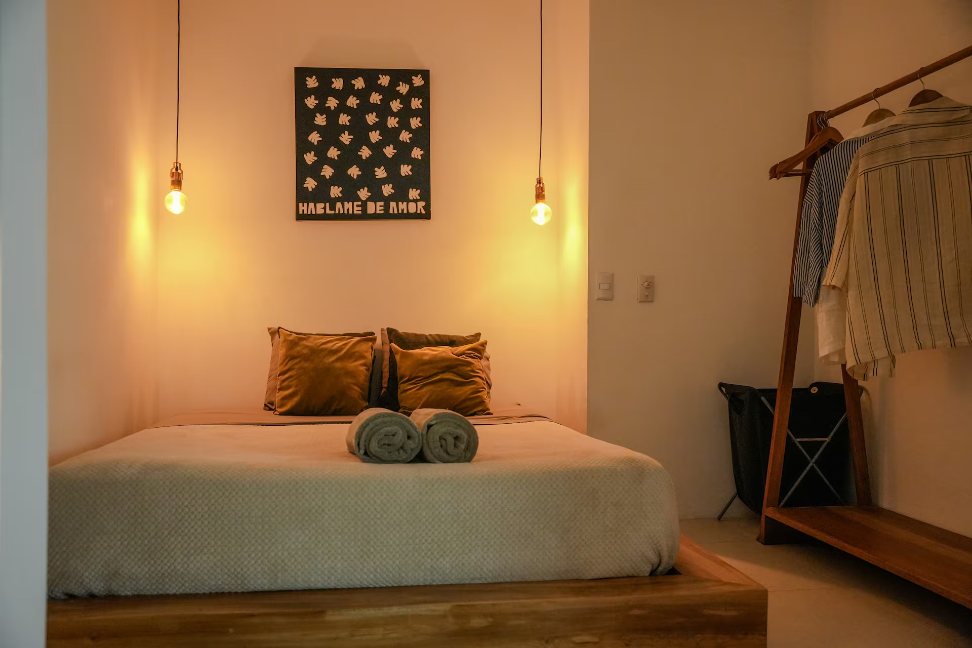 Bedroom with queen bed, mustard pillows, pendant lights, wooden clothing rack and minimalist decor