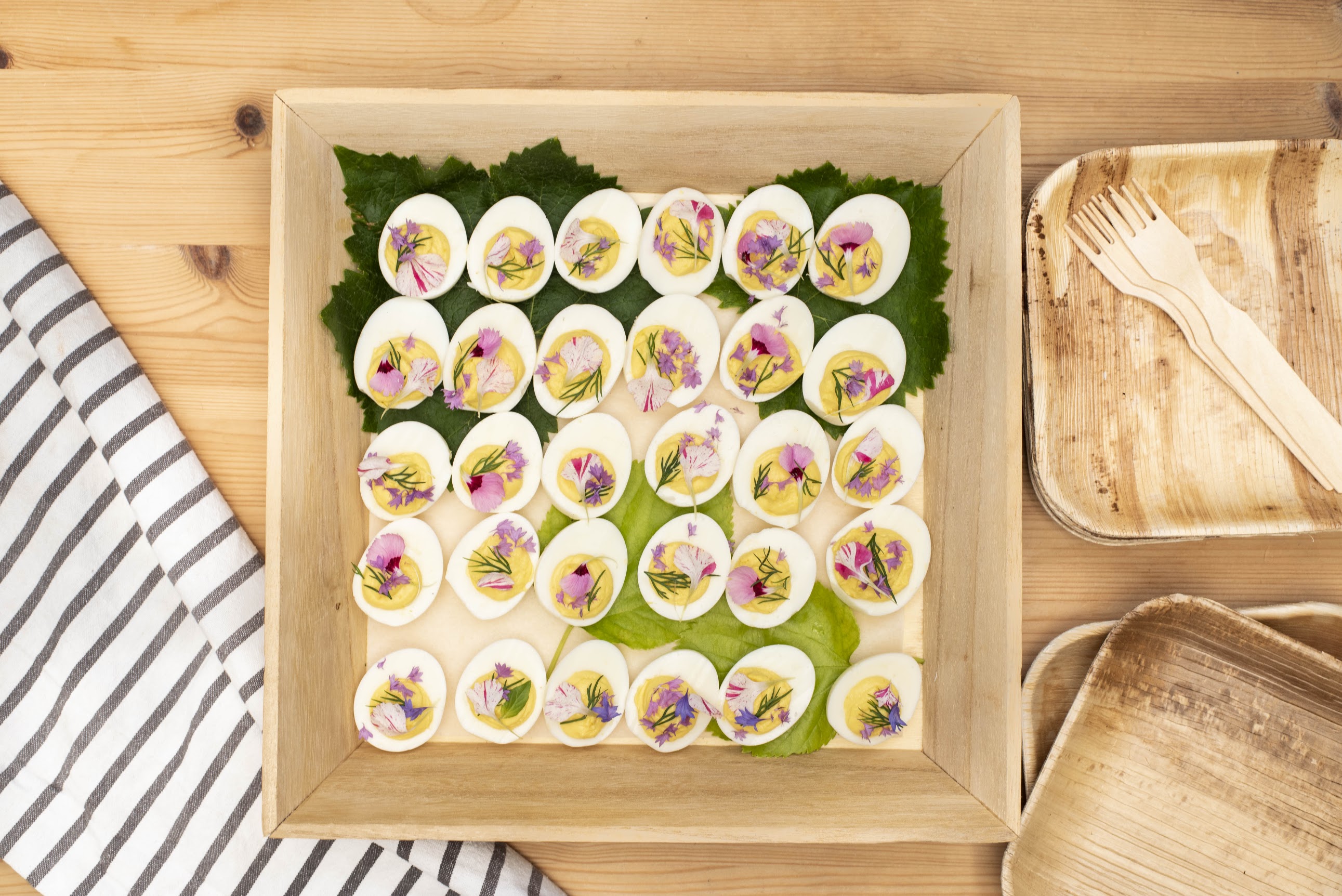 Just Fare grazing table deviled eggs garnished with edible flowers in a wooden tray with palm leaf plates