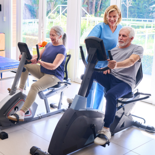An older couple pedaling on recumbent exercise bikes while a smiling clinician in blue scrubs supervises their cardiac rehab session