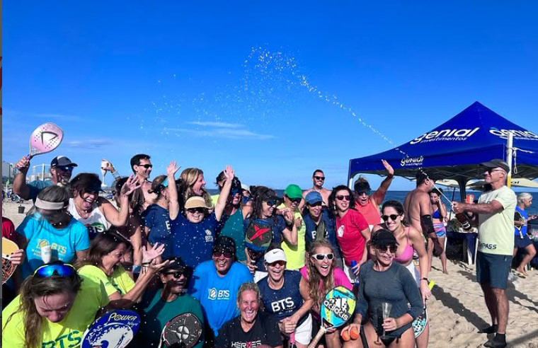 Grupo comemorando na praia com a tenda da Genial ao fundo