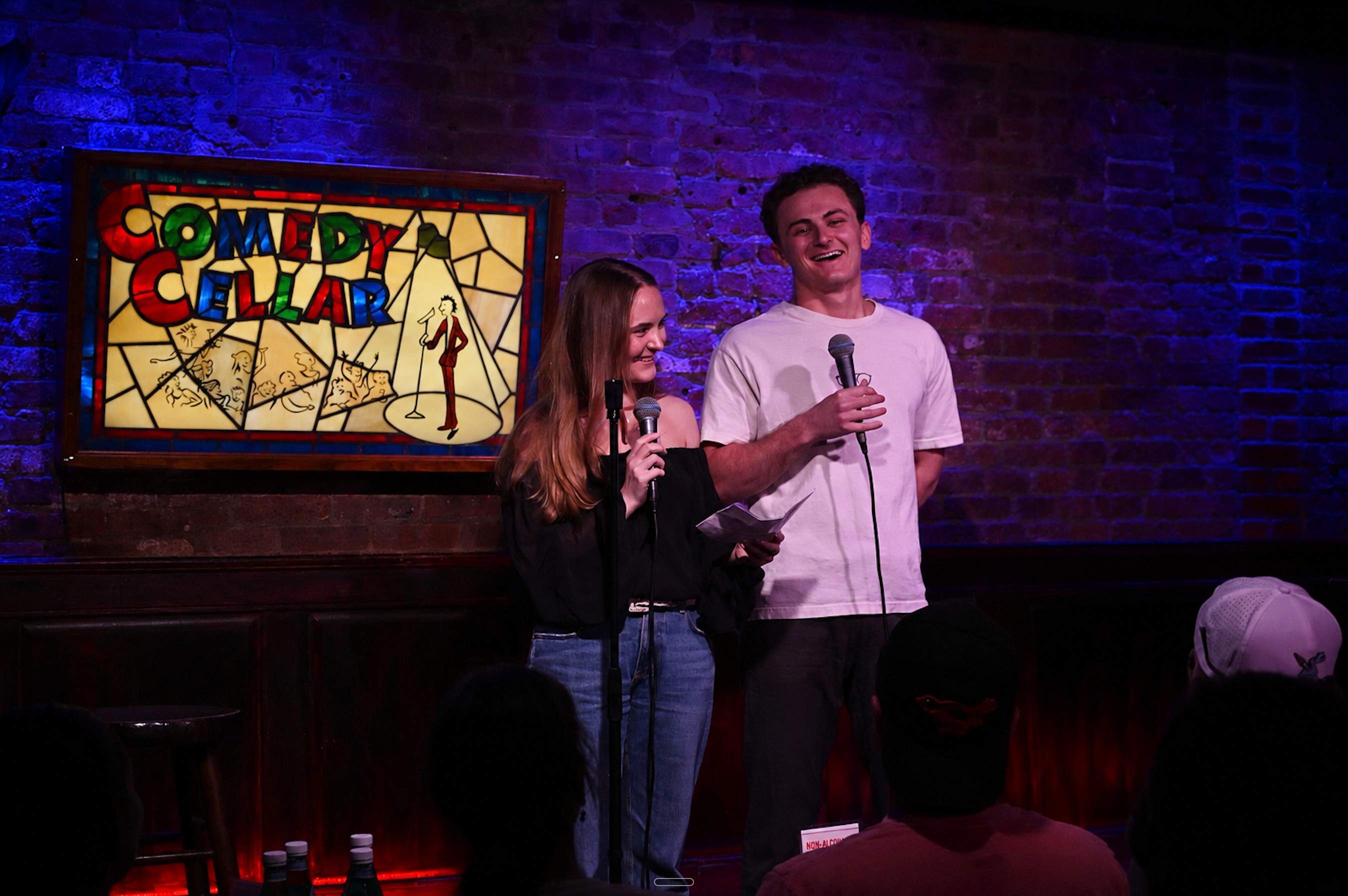 Evan and Marlee Golinsky on stage at the Comedy Cellar, holding microphones and laughing