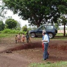 Europäischer Besucher und Kinder auf roter Erde vor einem Geländewagen unter Bäumen