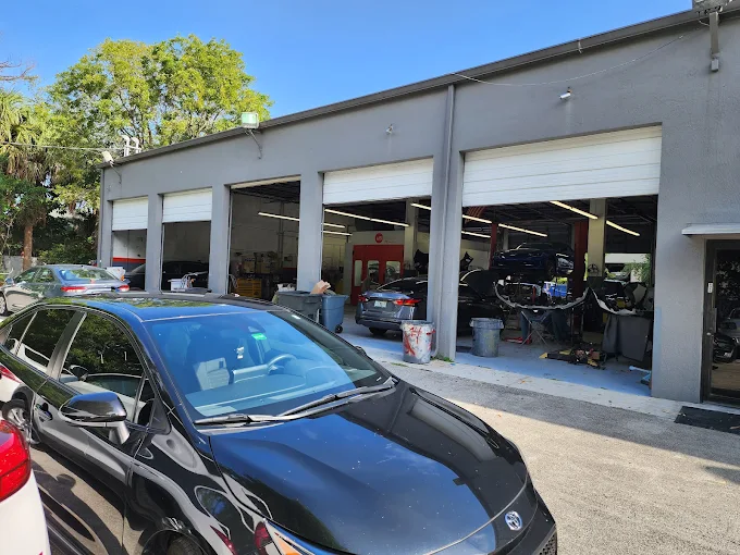 Wide exterior of Factory Finish Paint and Body shop with five bay doors open and vehicles being serviced in Lake Worth Beach, Florida