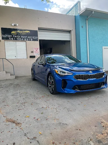 Blue Kia Stinger parked in front of the Factory Finish Paint and Body shop signage in Lake Worth Beach