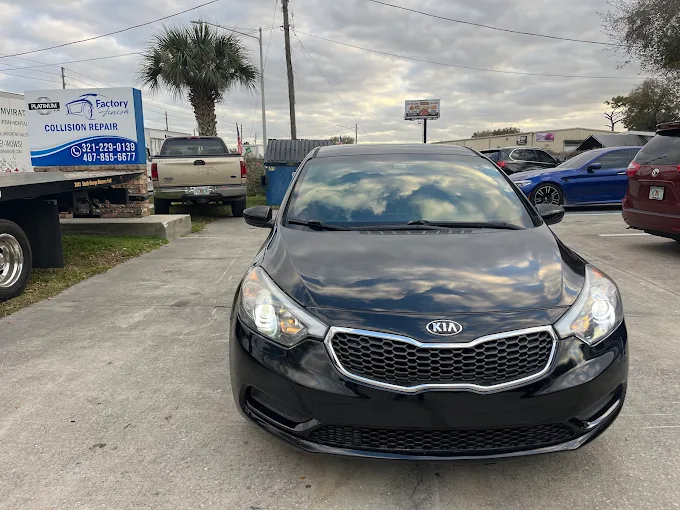 Black Kia sedan parked in front of a Factory Finish Collision Repair sign