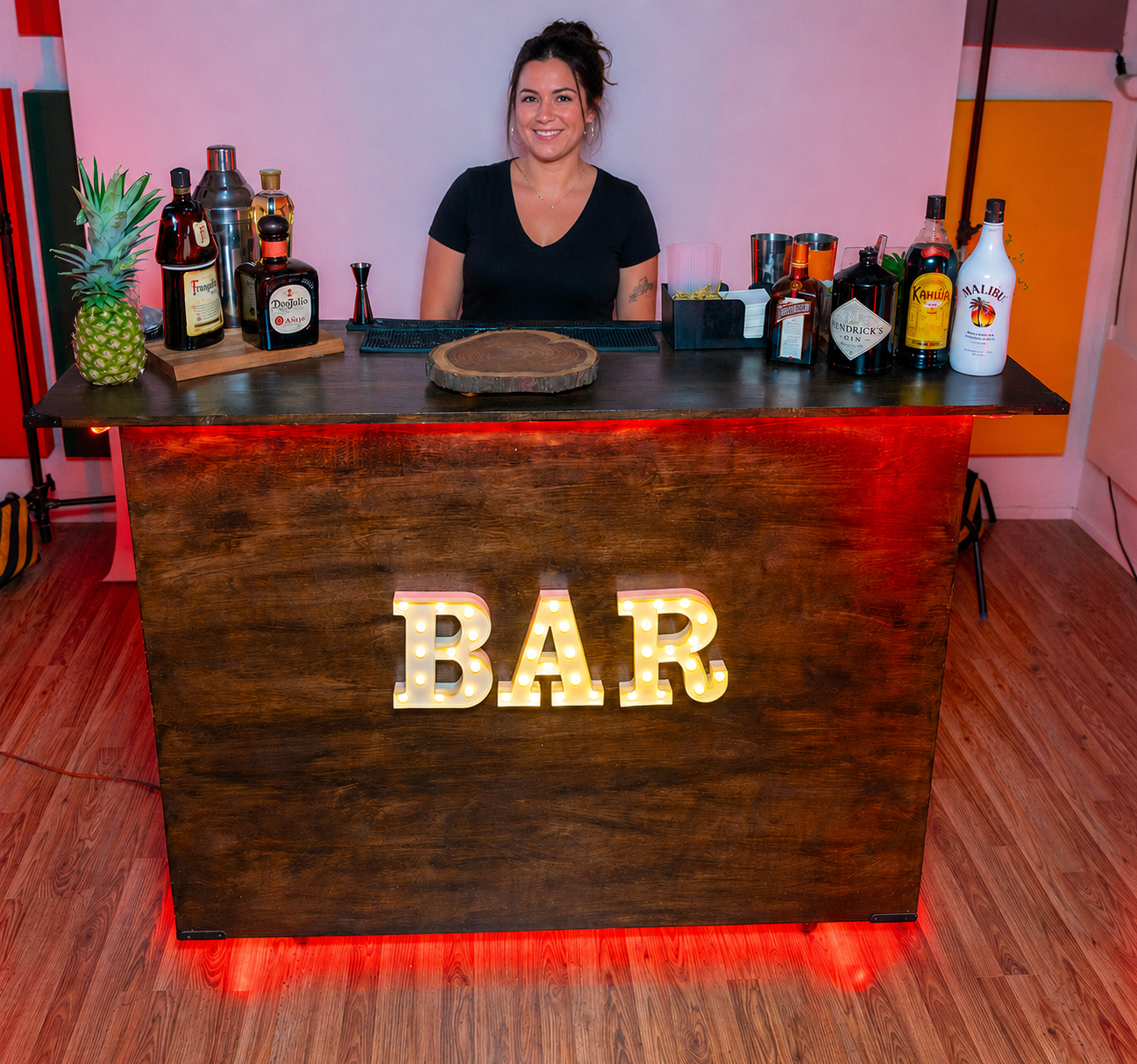 Professional female bartender smiling behind rustic marquee bar with red LED lighting - Fire Water Bars mobile bartending service South Florida