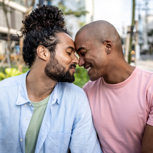 Two men sitting close together outdoors, smiling with their foreheads touching