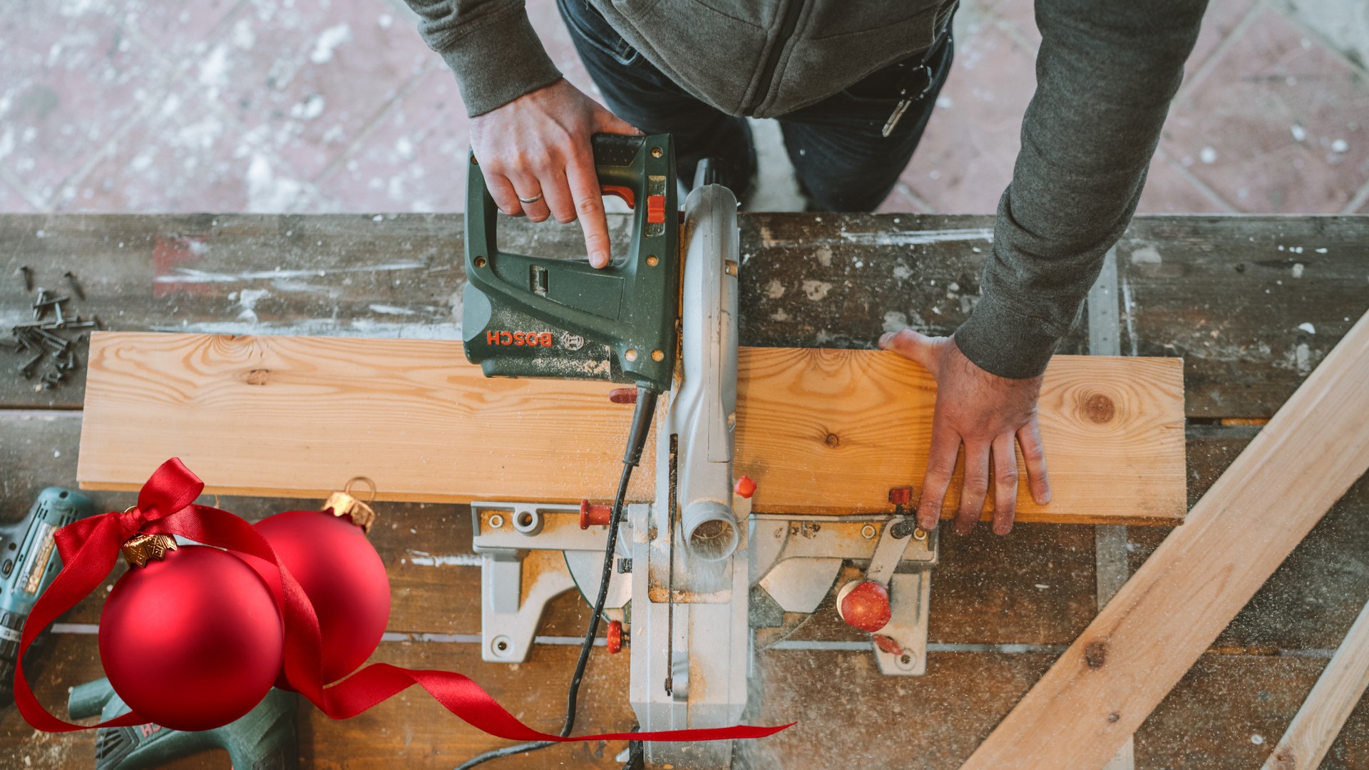 Overhead view of a contractor using a power saw to cut a wood board on a workbench, with red Christmas ornaments nearby