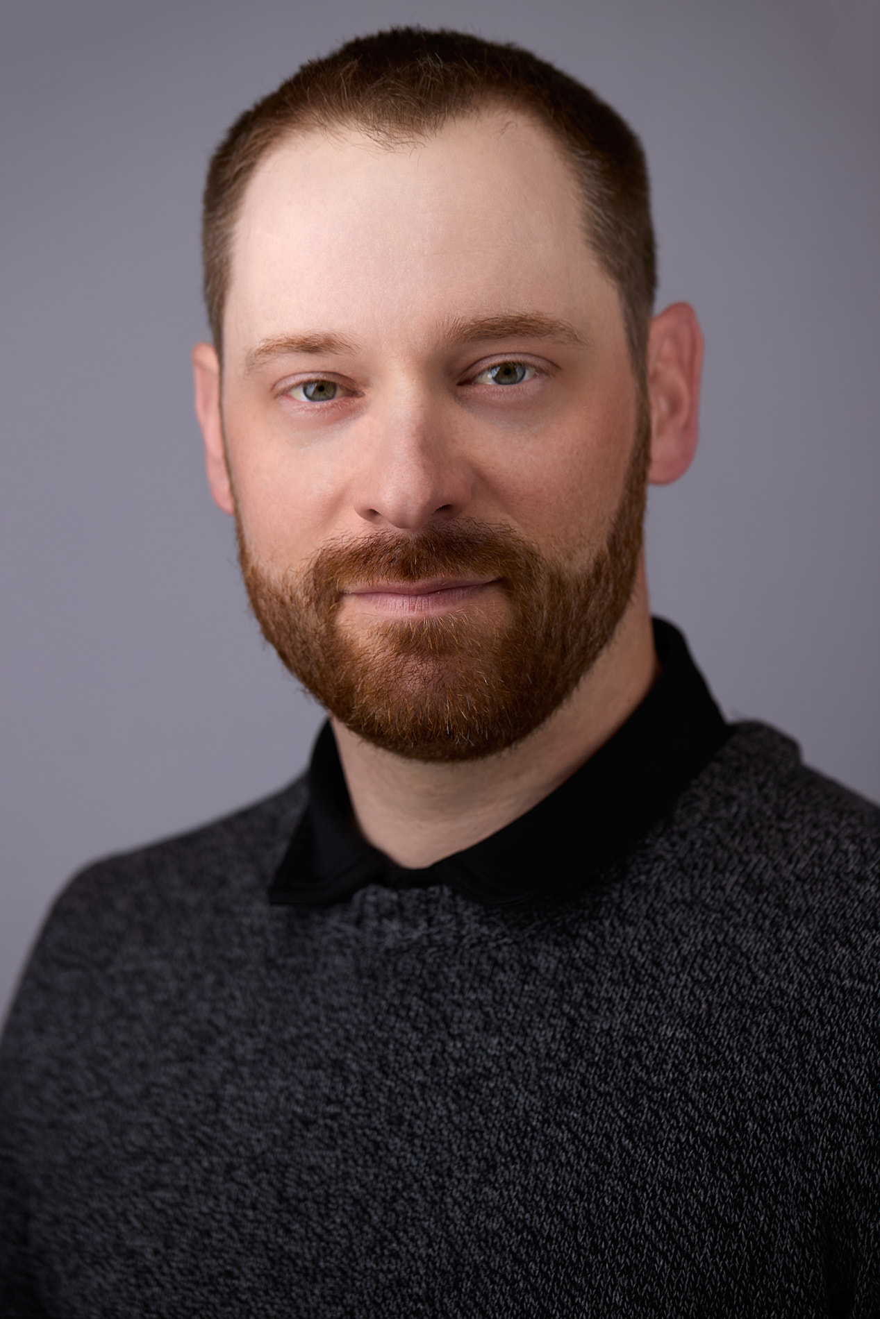 Professional headshot of man with reddish-brown hair and beard in dark collared shirt against gray background