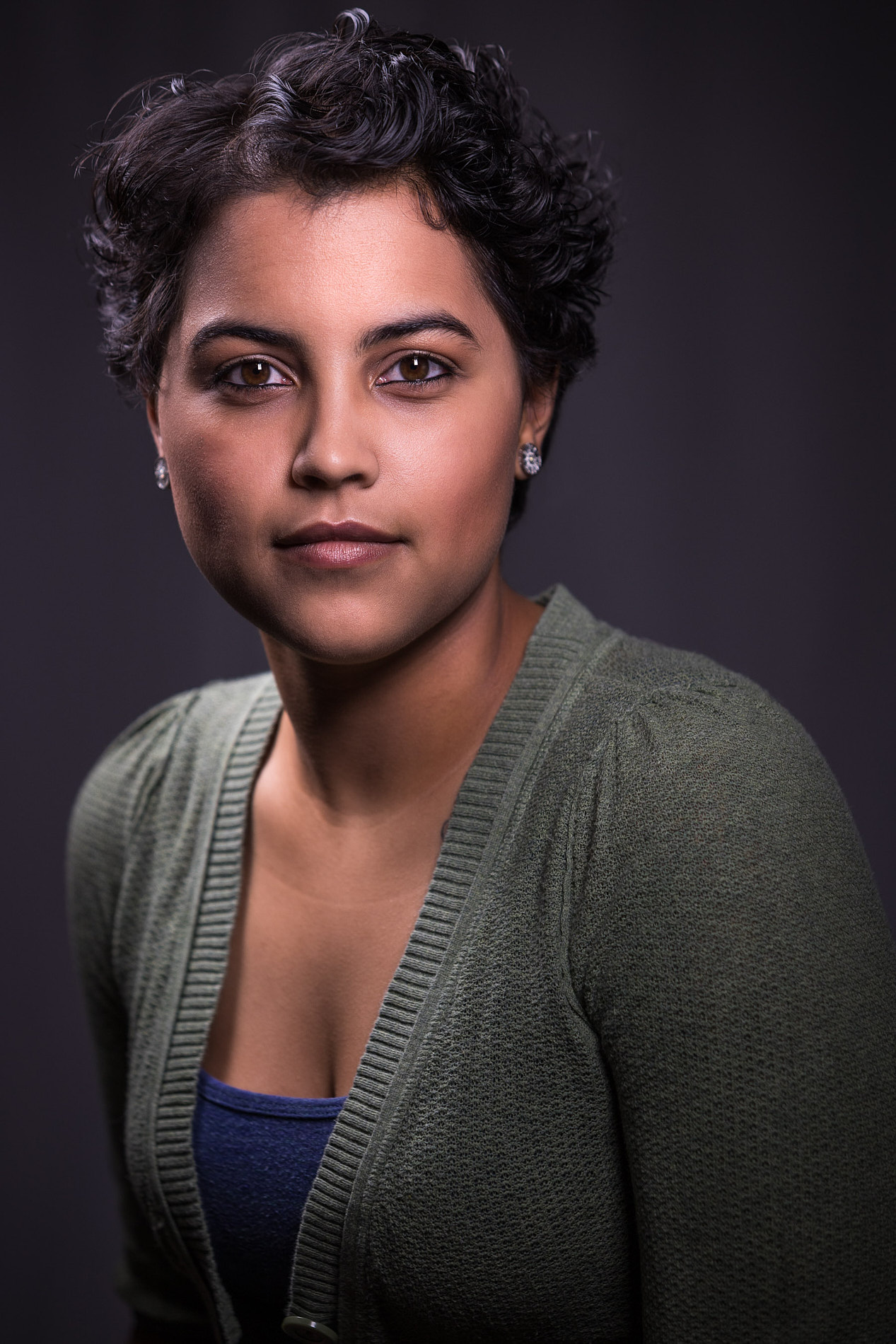 Professional headshot of woman with short curly dark hair in green cardigan over blue top against dark background