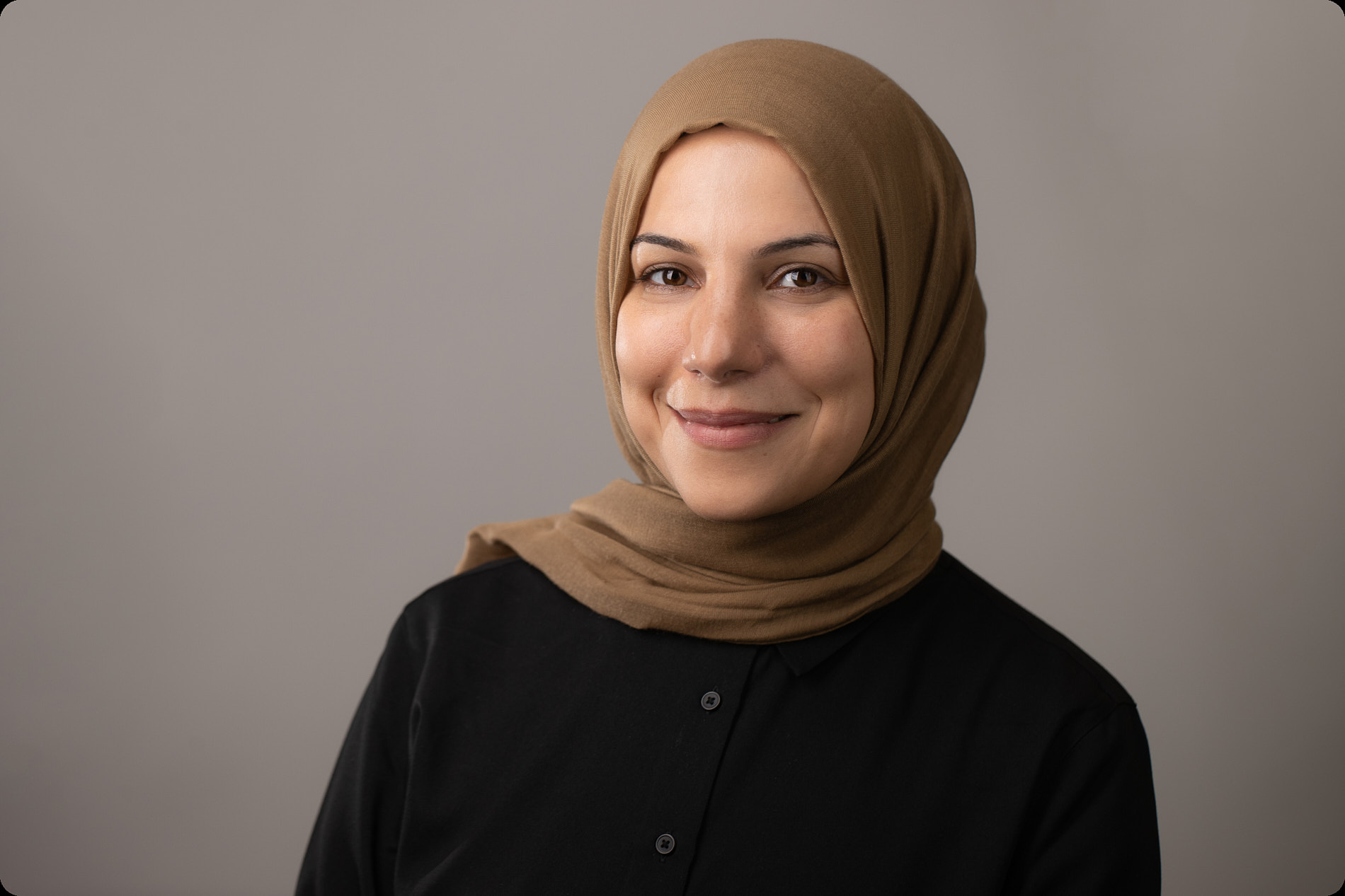 Professional headshot of woman wearing a tan hijab and black button-up shirt against a neutral background