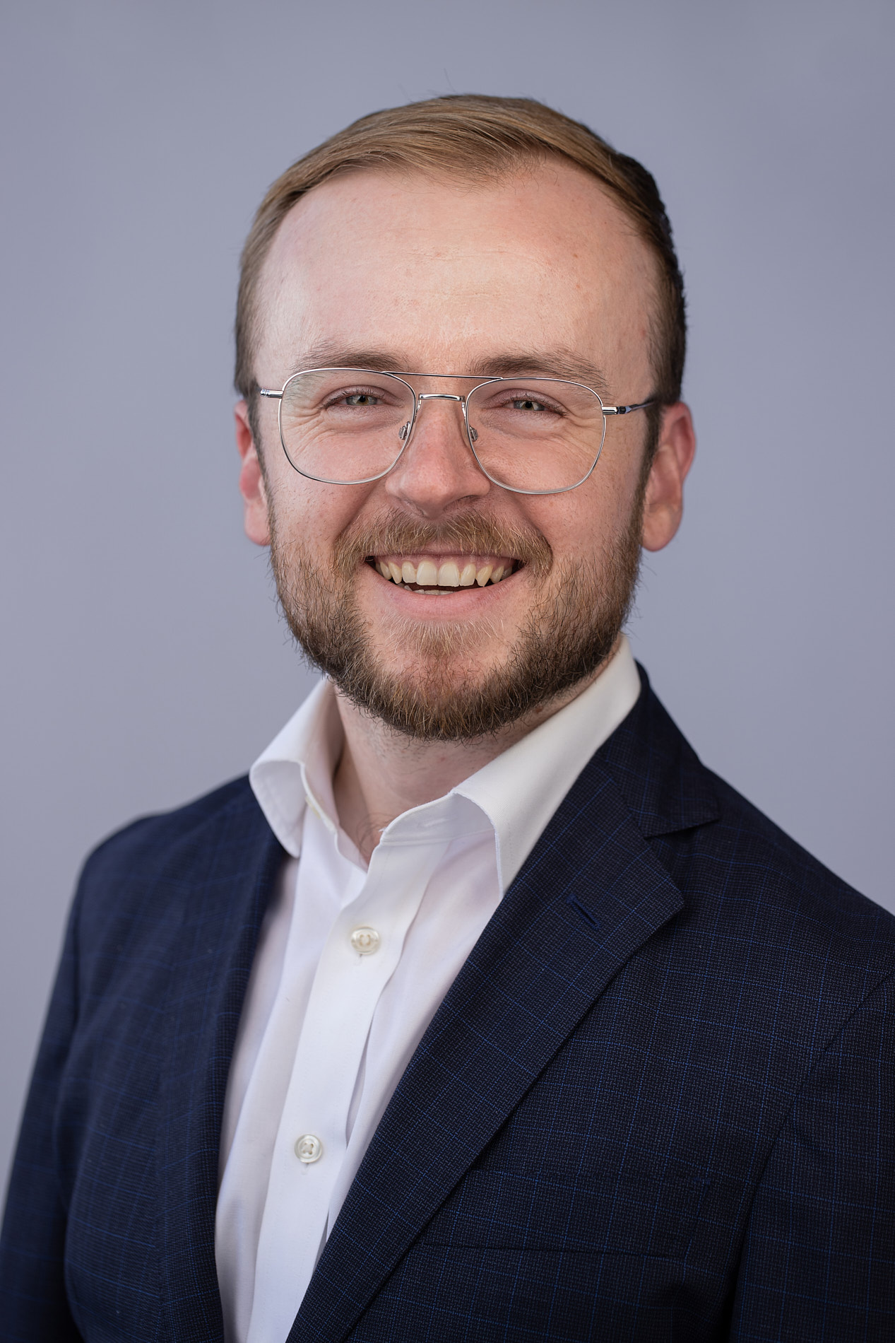 Professional headshot of man with light brown hair, beard and glasses in navy suit and white shirt against gray background