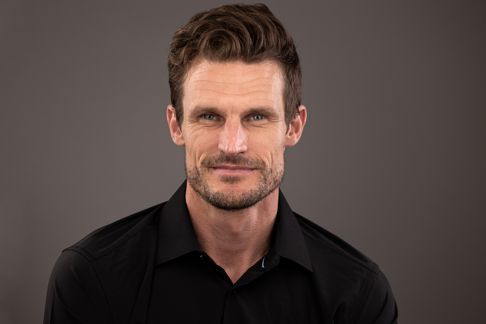 Professional headshot of man with brown hair and beard in a black shirt against a gray background