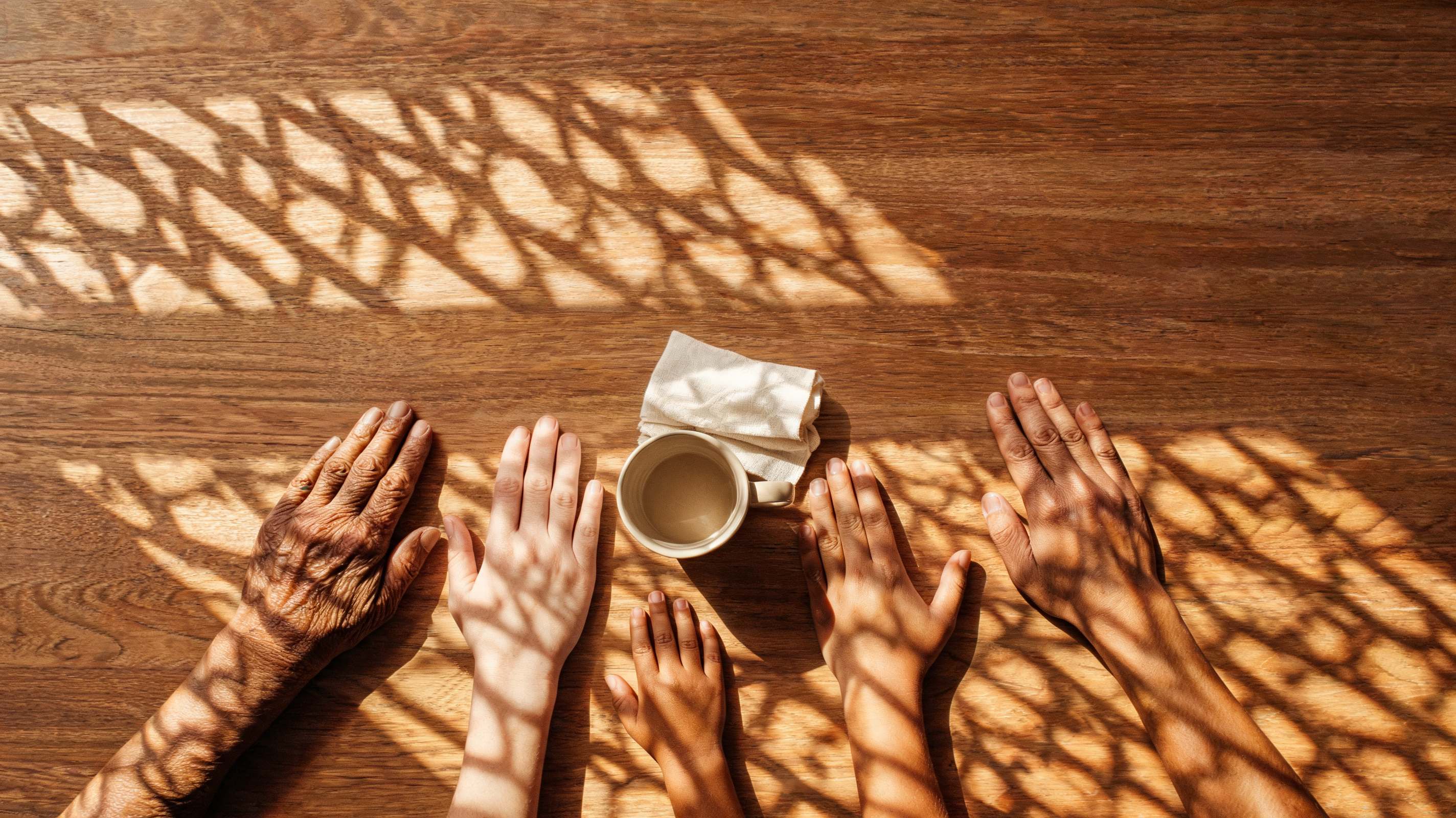 A warm overhead view of hands of three generations resting on a wooden table around a shared cup, dappled daylight