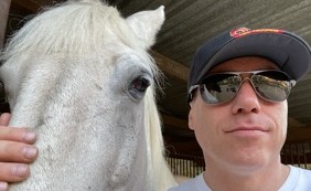 Ian King wearing a cap and sunglasses beside a white horse at a stable.