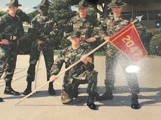 Marine recruits in camouflage uniforms holding a unit guidon flag.