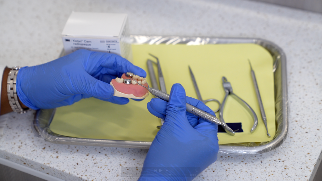 Gloved hands working on a typodont with a dental instrument beside a tray of tools and Ketac Cem cement