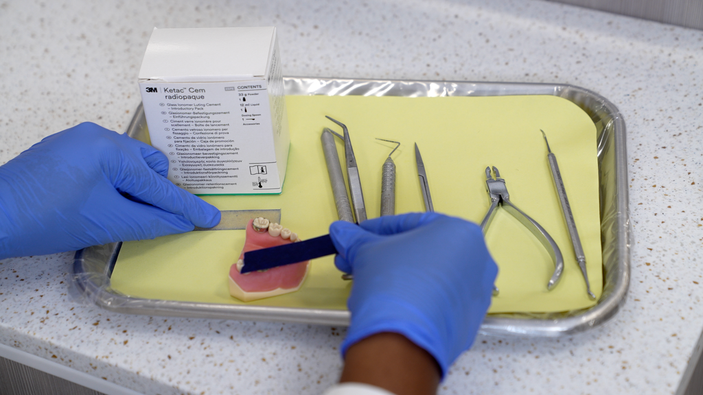 Gloved hands holding a typodont and spatula next to a tray with Ketac Cem glass ionomer cement and instruments