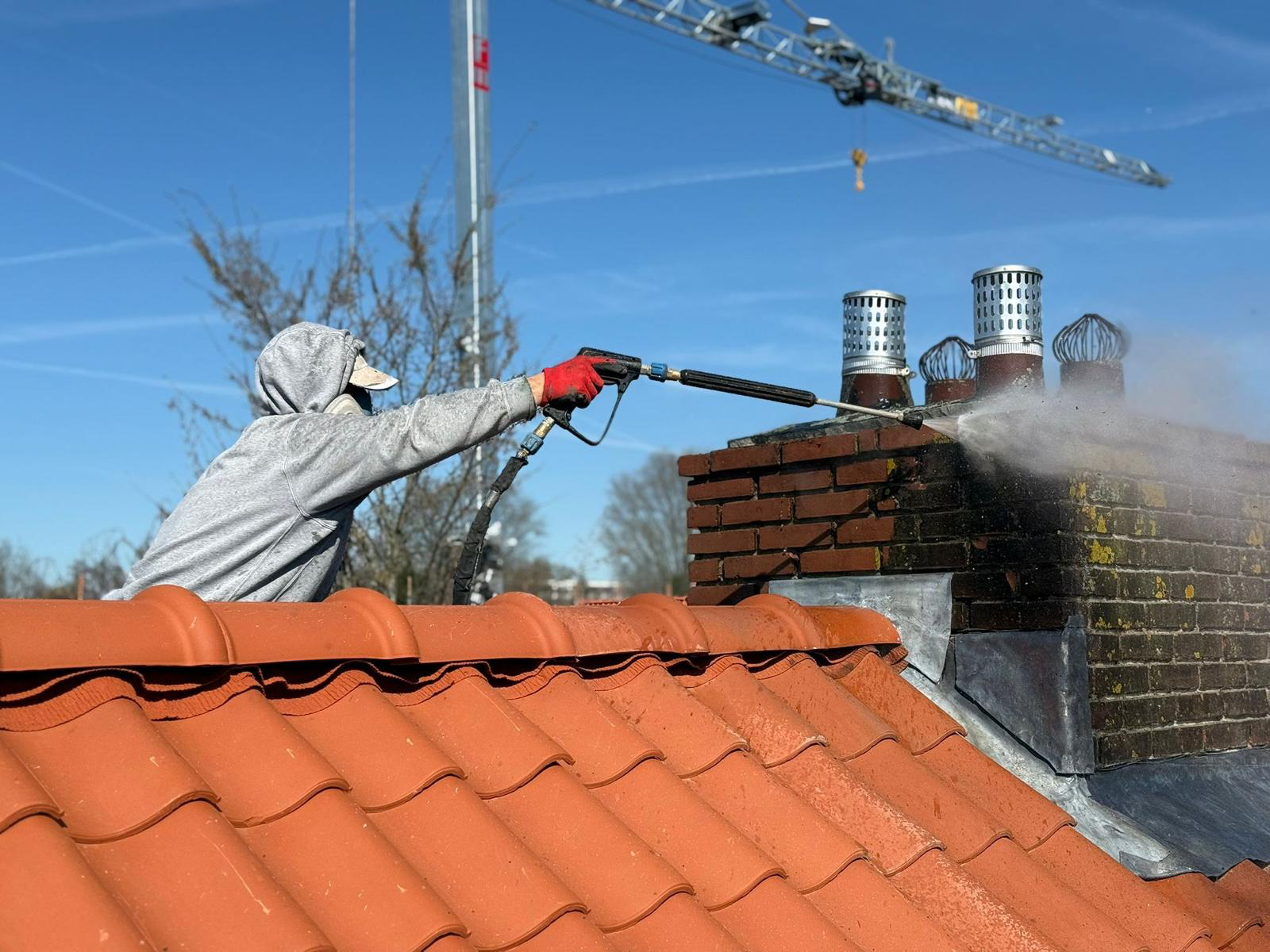 Renovox medewerker stoomt schoorsteen schoon op terracotta dak — blauwe lucht met bouwkraan