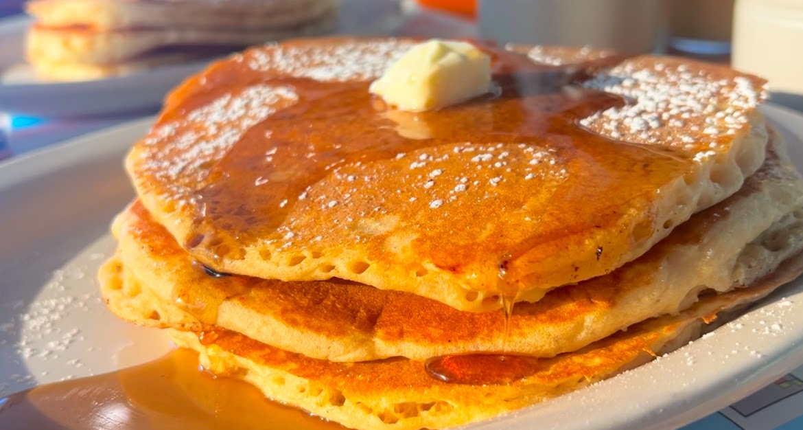 A tall stack of pancakes with syrup, fresh strawberries, and whipped cream