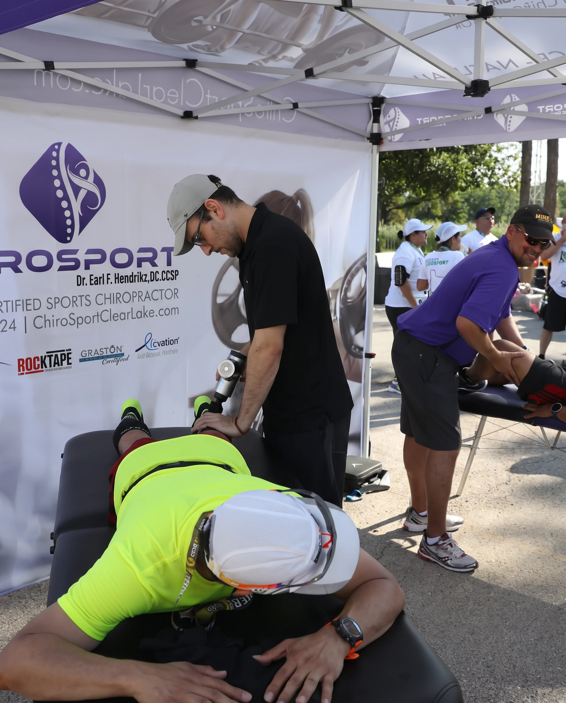 A practitioner using a percussion massage tool on a runner's calf at a road race