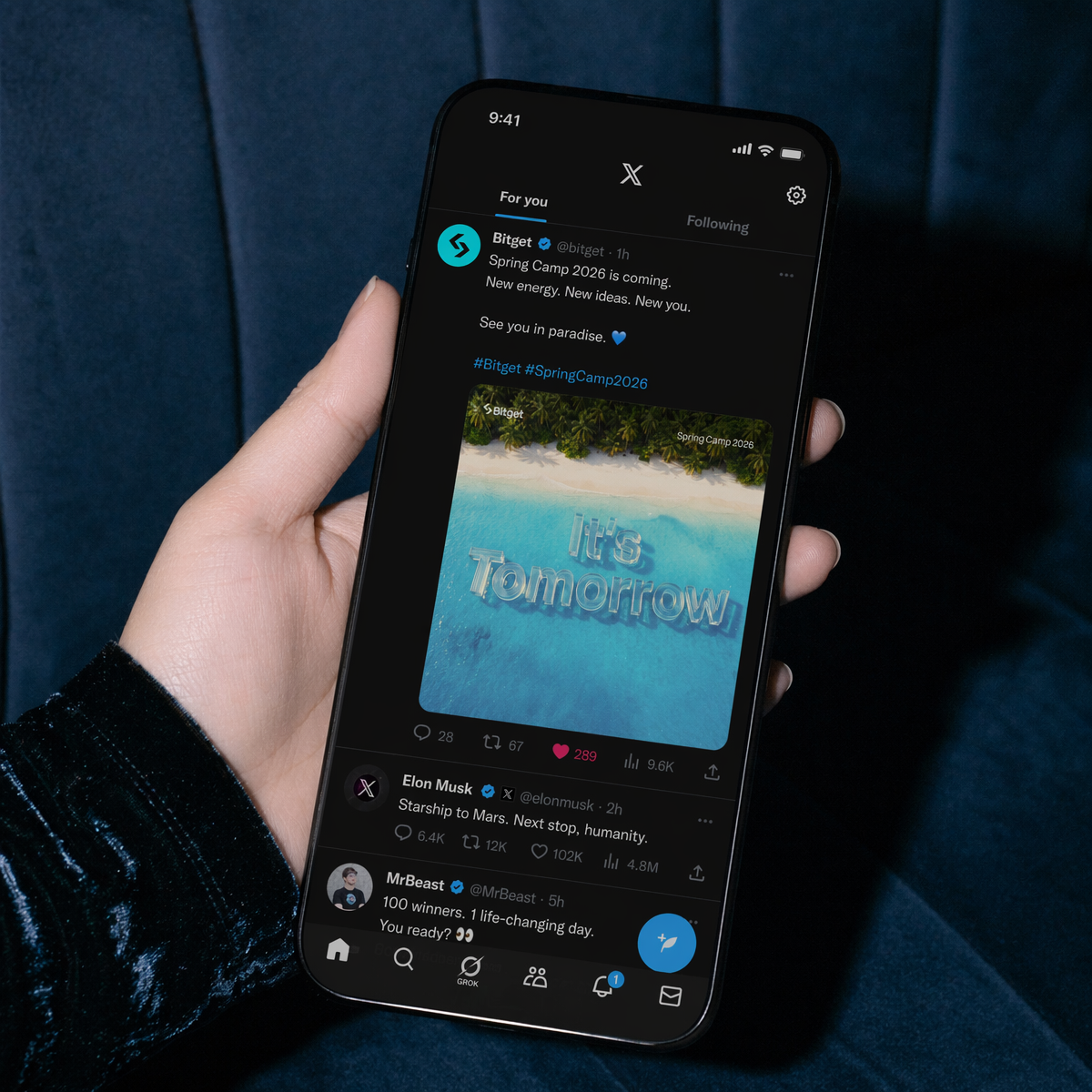 Bitget Spring Camp 2026 social — hand holding iPhone showing Bitget X/Twitter post with It's Tomorrow beach teaser