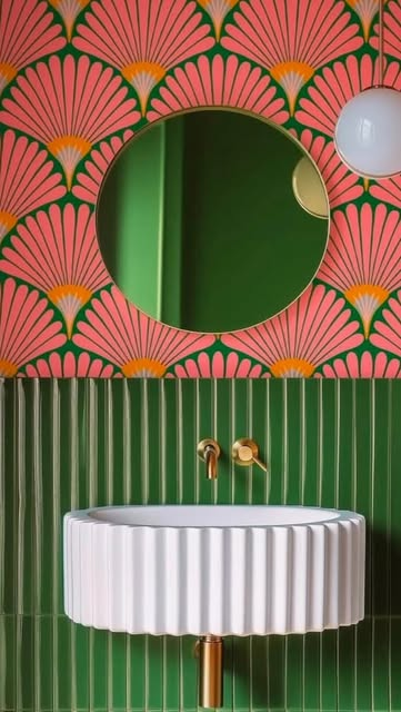 Bold bathroom with coral pink art deco fan-pattern wallpaper, forest green paneling, white fluted cylindrical basin on brass pedestal, and circular green mirror