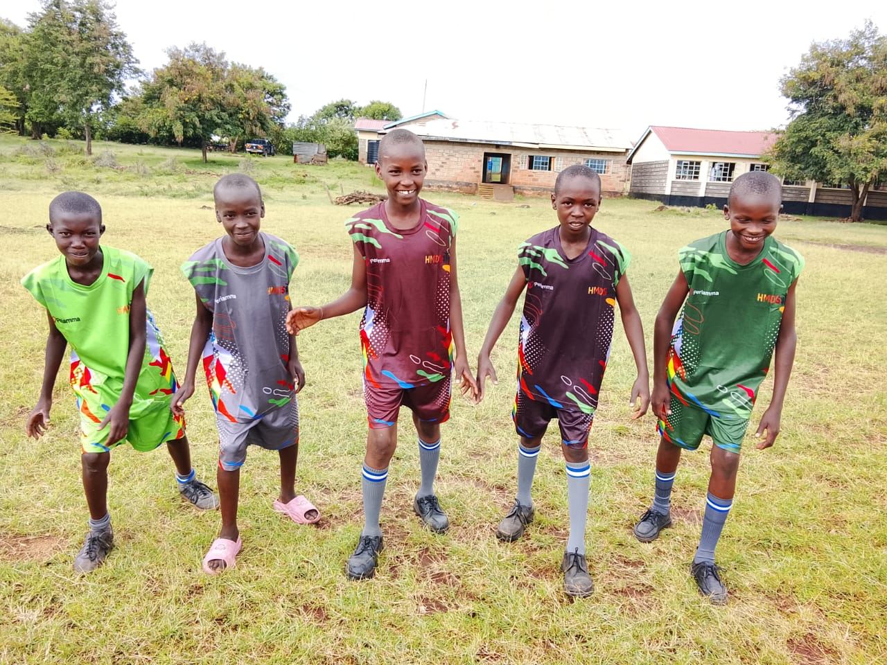 Five Kenyan children in colorful Periamma athletic jerseys standing on a grass field
