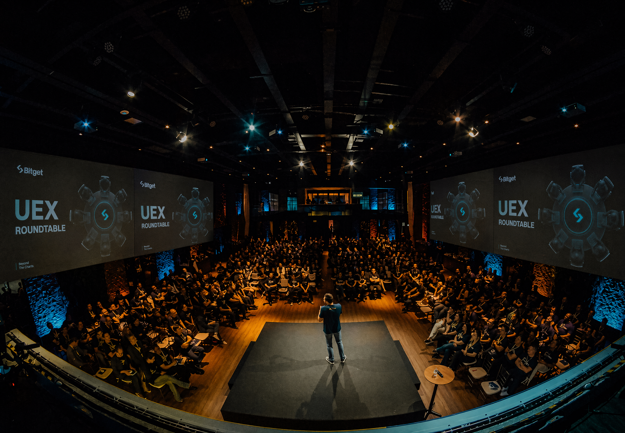 Bitget UEX Roundtable — fisheye shot of packed live event venue with four branded screens flanking the stage