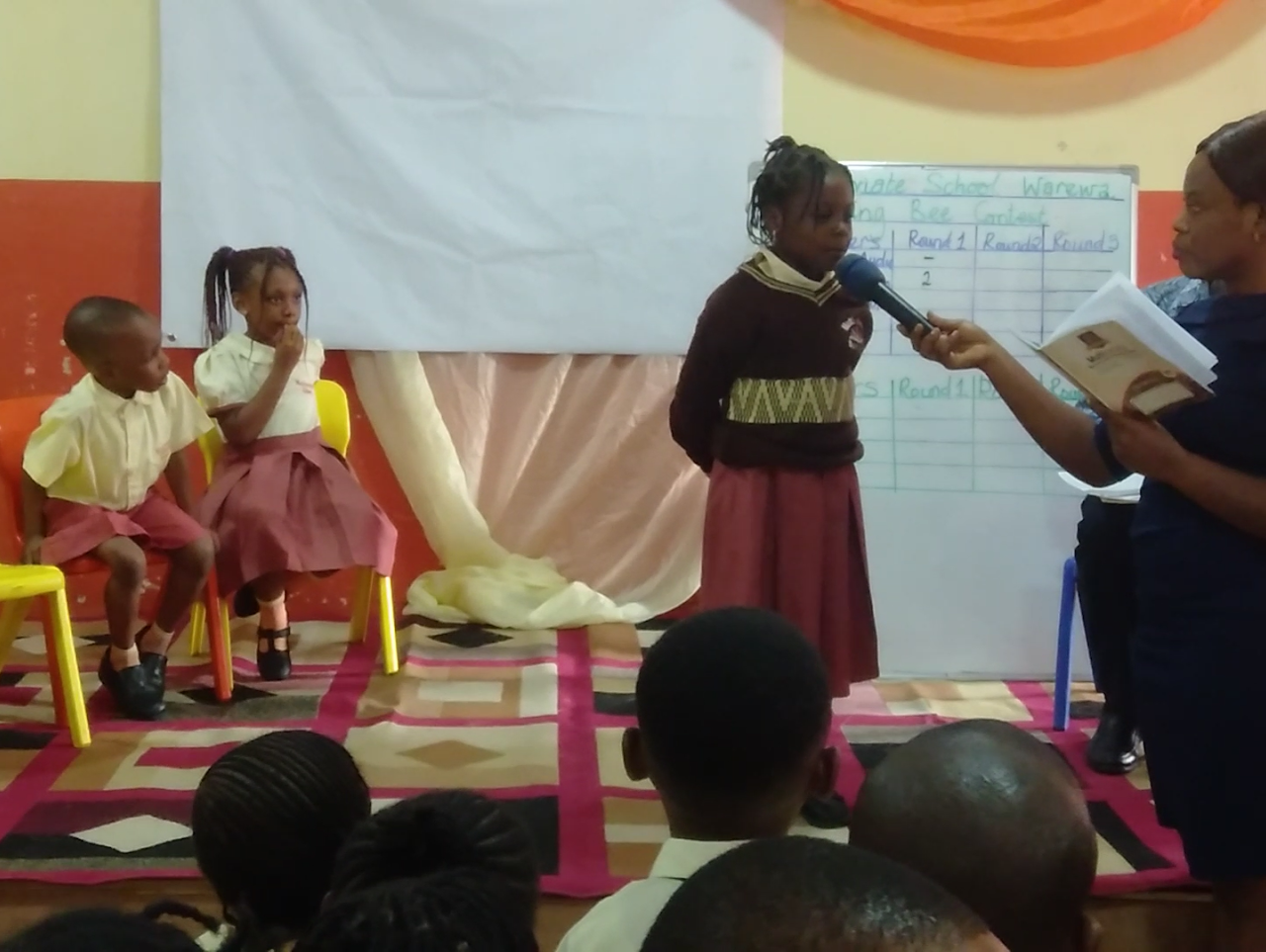 Children on stage during a spelling bee contest with a scoring whiteboard