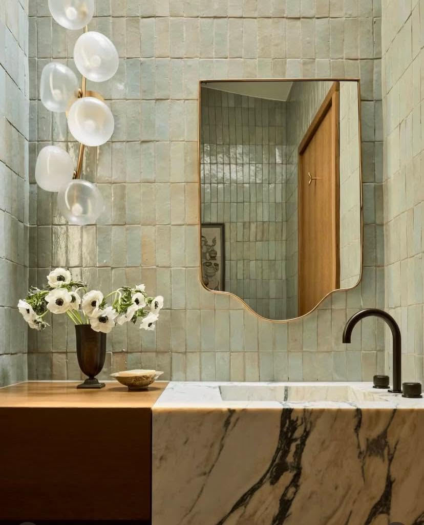 Elegant bathroom with sage-green gridded tiles, white frosted globe pendants, curved brass mirror, and marble countertop with white flowers — bathroom design by Jayne Michelle Interiors