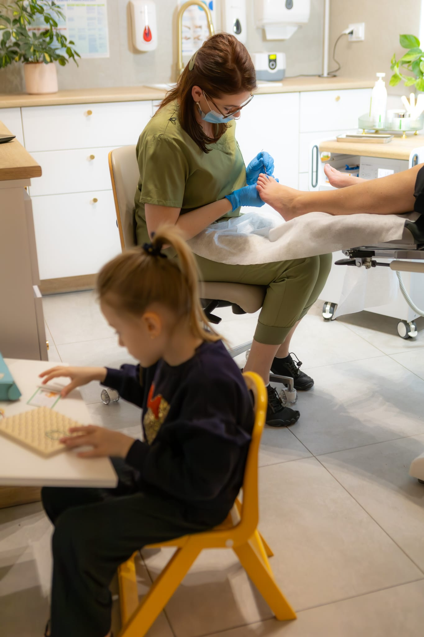 Karolina Szynwelska w żółto-zielonym stroju wykonująca zabieg podologiczny na stopie pacjenta, w tle dziewczynka siedząca na żółtym krześle