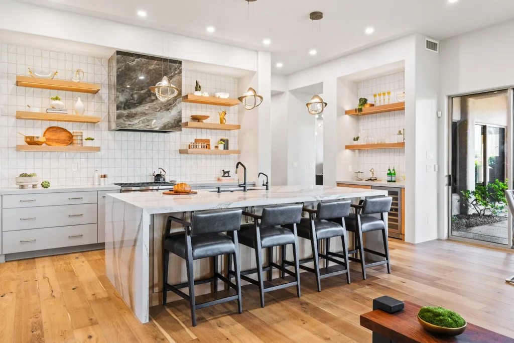Modern white kitchen with marble waterfall island, leather barstools, floating wood shelves and grid tile backsplash