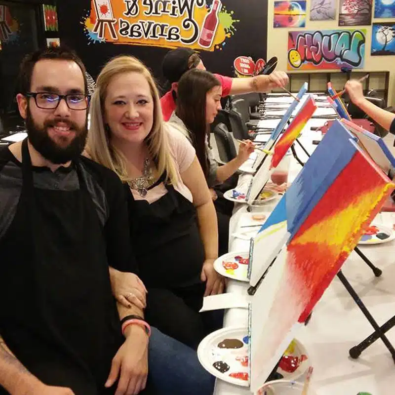 Couple posing at Wine & Canvas with colorful paintings in progress behind them