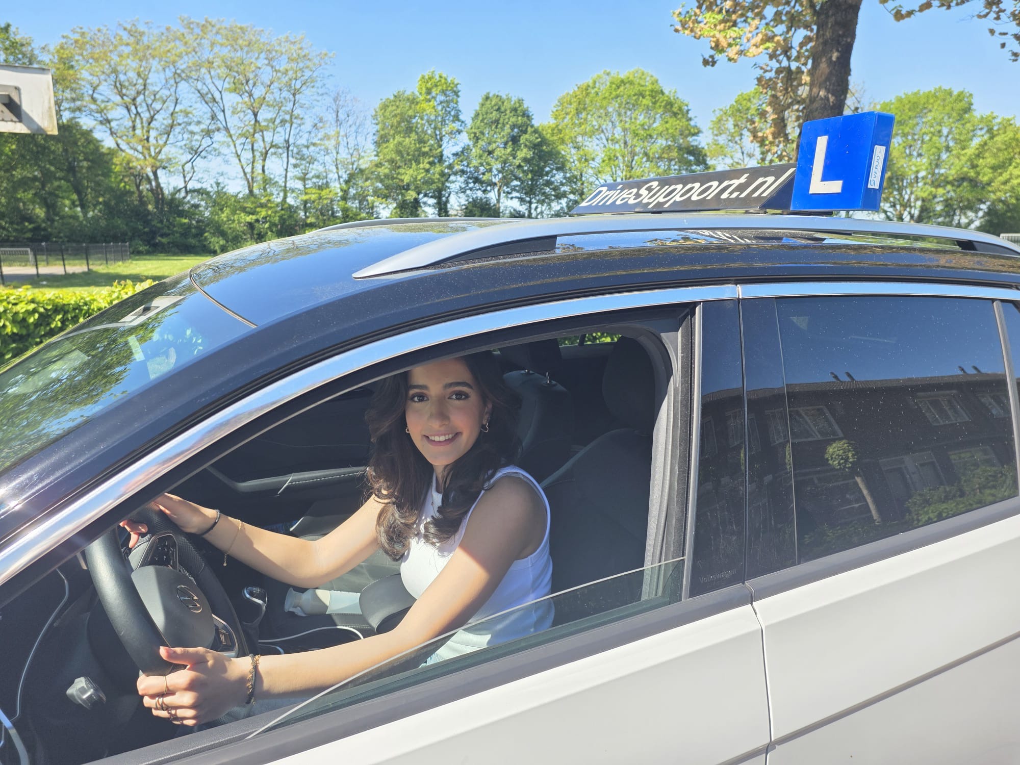 Leerling zit glimlachend achter het stuur van de DriveSupport lesauto op een zonnige dag