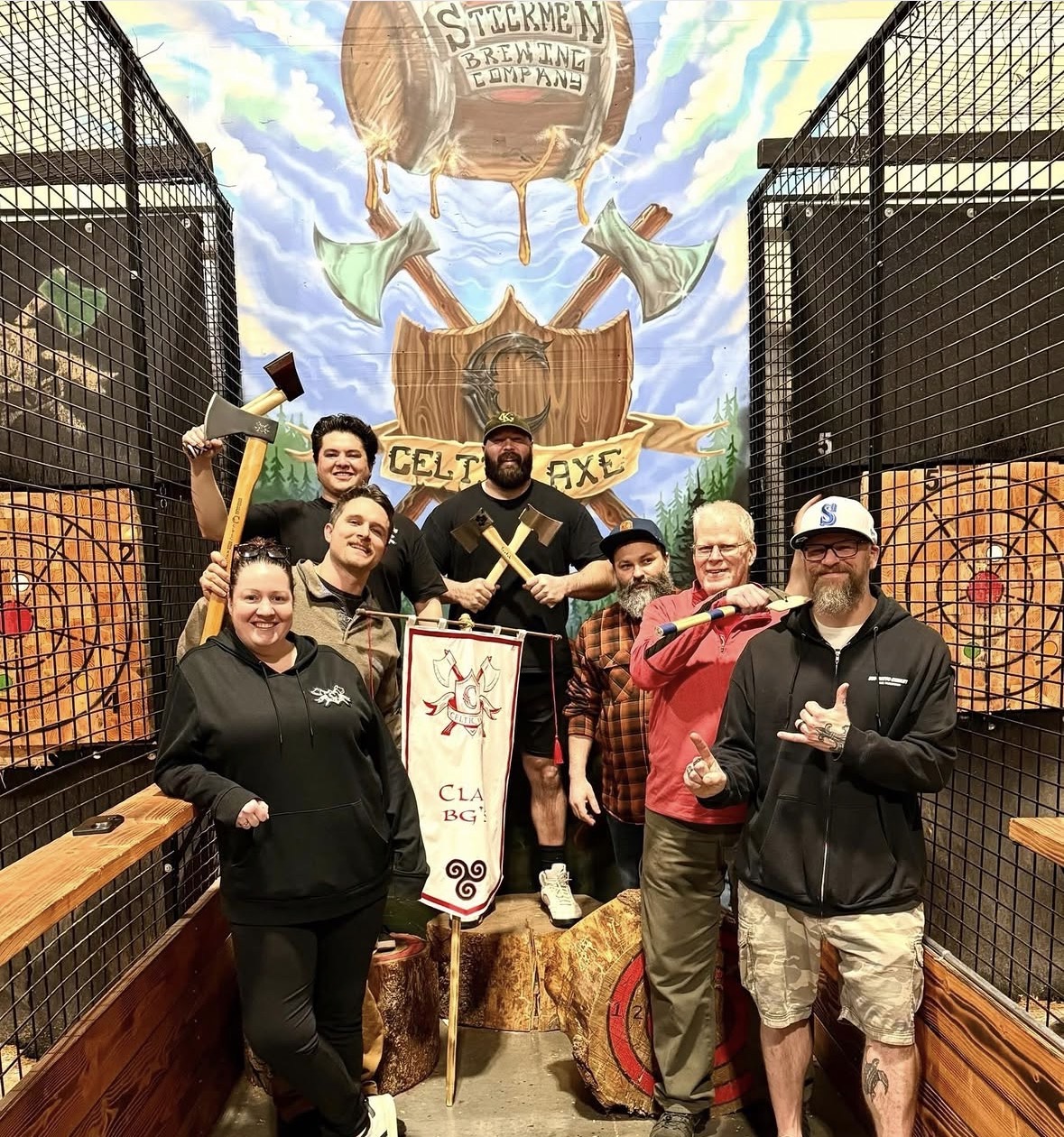 Group of seven people posing with axes and a Celtic Axe clan banner in front of the Stickmen Brewing Company Celtic Axe mural