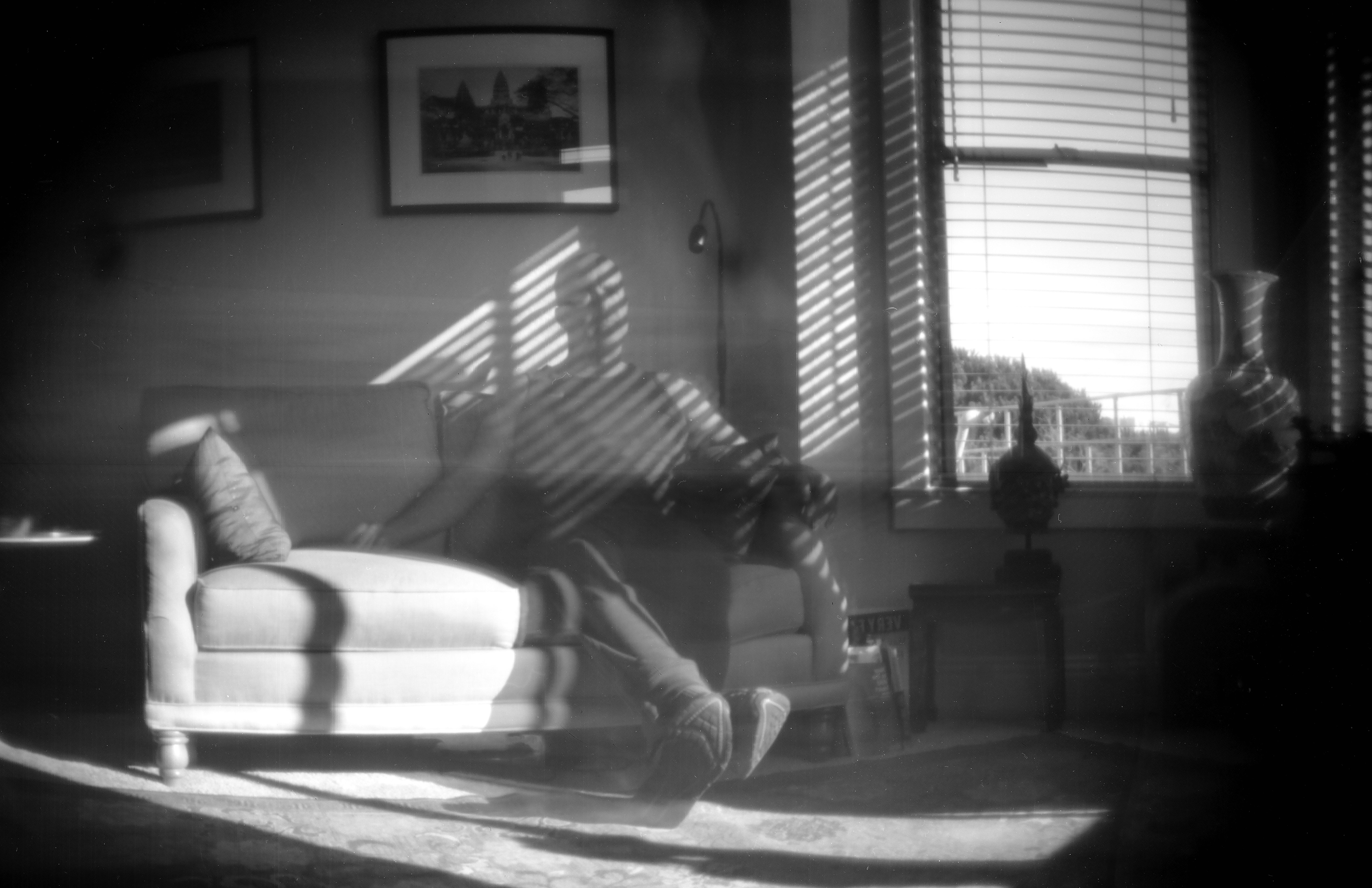 Ethereal pinhole photograph of living room interior with sofa and venetian blinds casting dramatic light stripes, ghostly motion blur figure