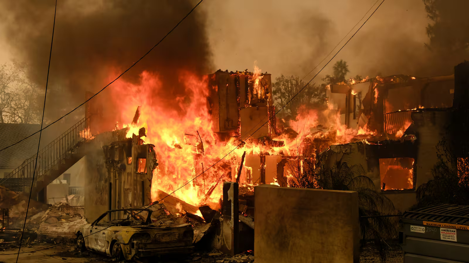 Building engulfed in flames during active wildfire with burned vehicle in foreground