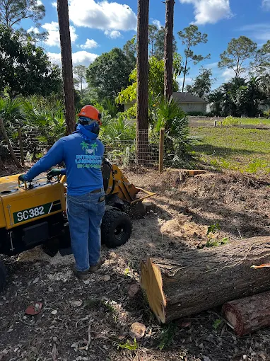 Integrity Landscape crew member in branded uniform operating a stump grinder beside freshly cut pine logs