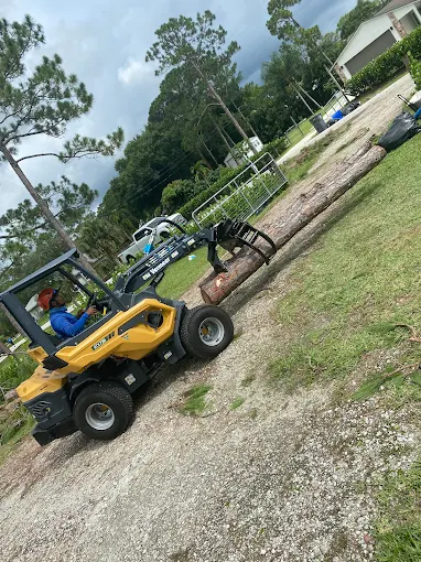 Yellow skid-steer machine hauling a long pine log across a cleared property during debris cleanup