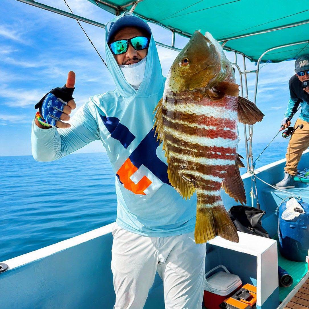 Jeffrey Chavarría, con ropa de pesca azul, sostiene un pez rayado a bordo de una embarcación bajo una cubierta turquesa.
