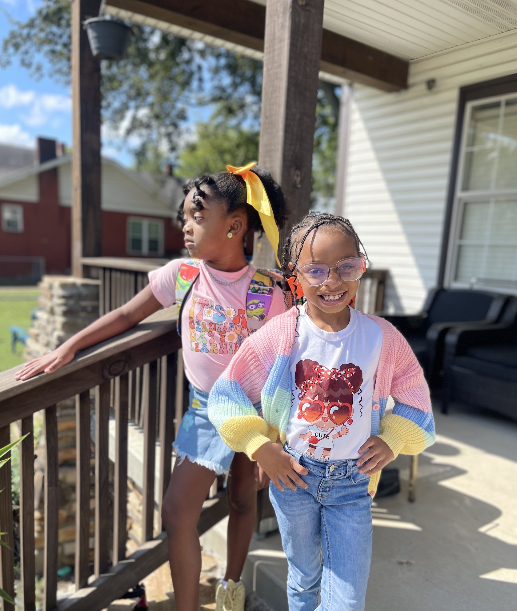 Two happy students smiling on a sunny porch - one with a yellow bow, the other in a colorful rainbow cardigan