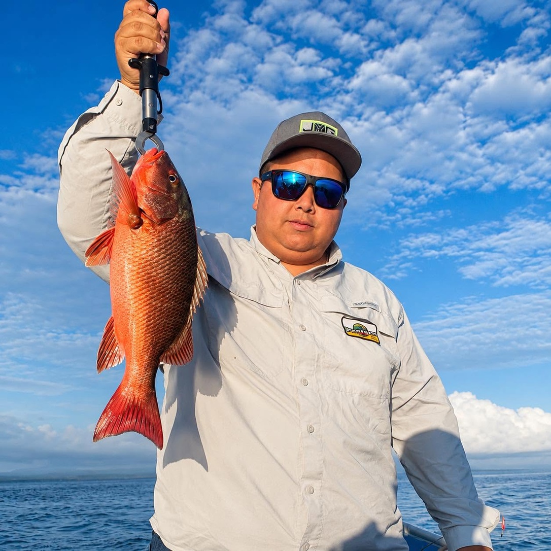 Leonardo Chavarría sostiene un pargo rojo a bordo de una embarcación, con el mar y el cielo despejado al fondo.
