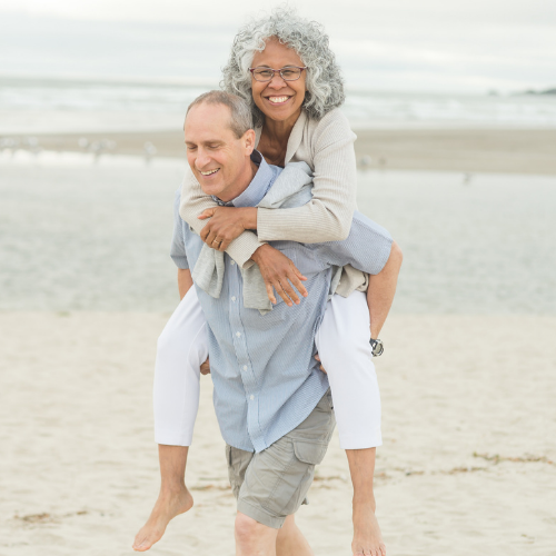 A smiling older man giving a laughing woman with curly gray hair a piggyback ride on the beach