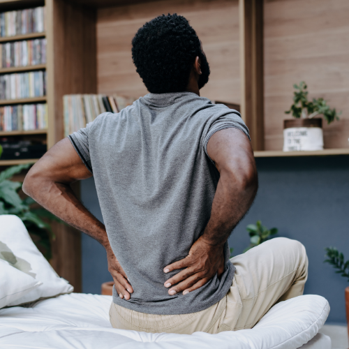 A man in a gray shirt sitting on the edge of a bed, seen from behind, holding his lower back in pain