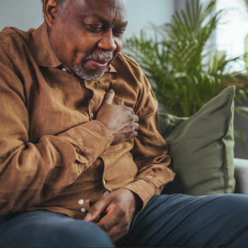 An older man in a brown shirt sitting on a couch with his hand pressed to his chest in discomfort