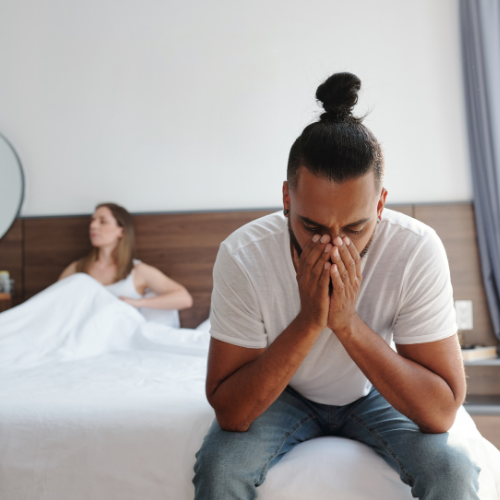 A man sitting on the edge of a bed with his face in his hands while his partner sits turned away in the background