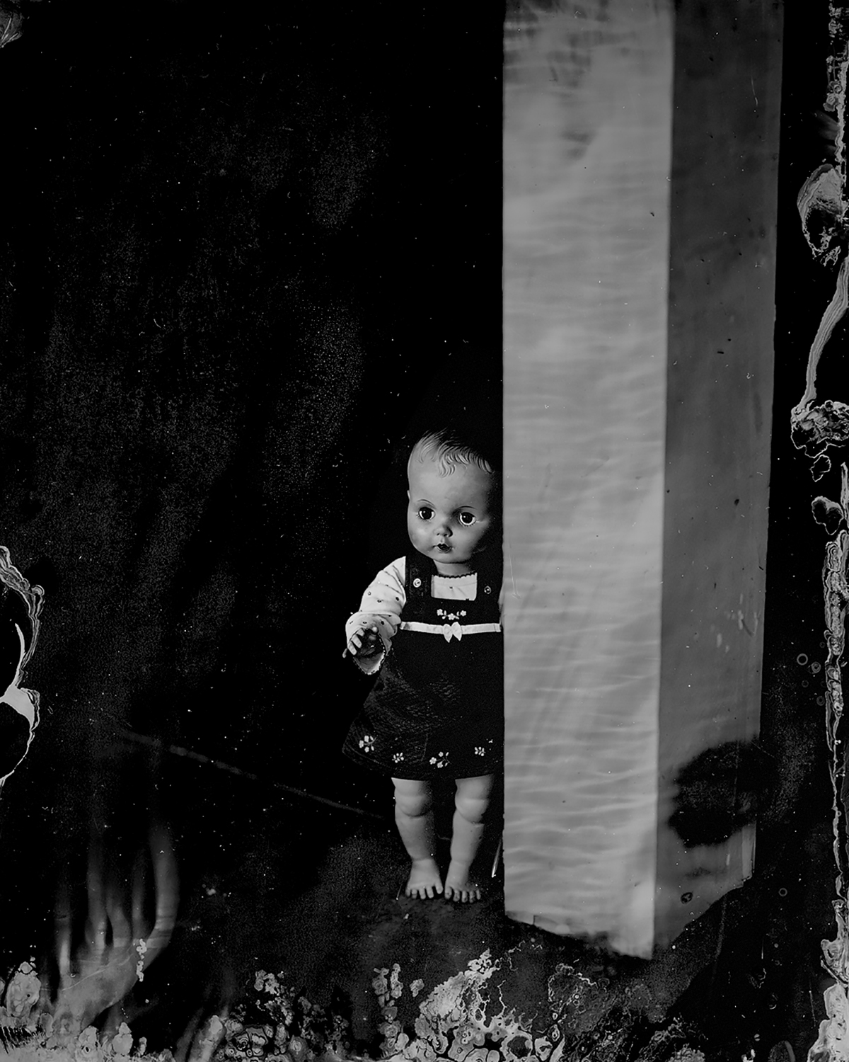 Haunting ambrotype of a vintage baby doll standing beside a white column against deep black background with chemical artifacts and emulsion textures along the edges