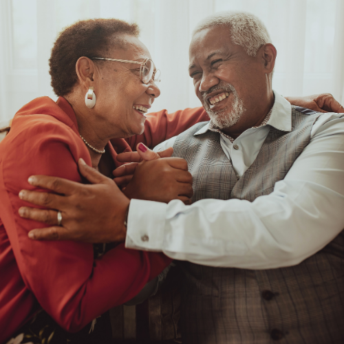 An older couple laughing together as they embrace, the woman in a red jacket and the man in a gray vest