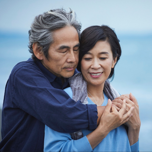An older man with gray hair hugging a smiling woman from behind by the ocean, their hands clasped together